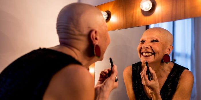A bald woman looks in the mirror while putting on red lip stick and smiling to herself.