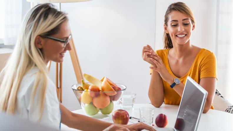 A dietician sits on one side of a desk with fruit and a laptop on it and a patient sits on the other side.