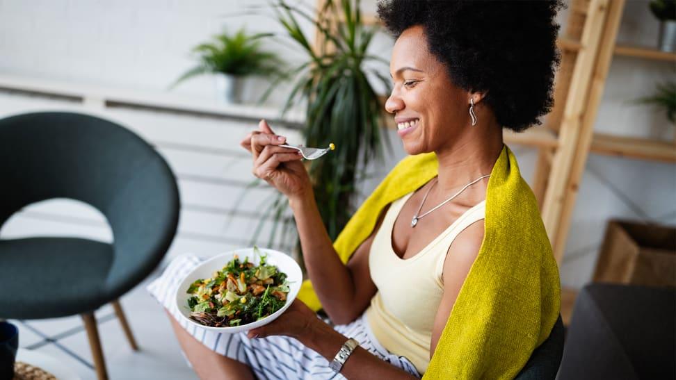 A person sits on a chair and holds a bowl with salad in it while smiling.