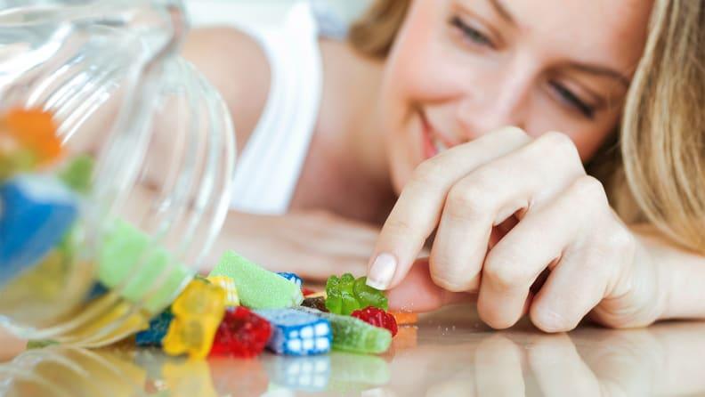 A candy jar tipped over to have candy spilling out of it and a woman reaches for a piece.
